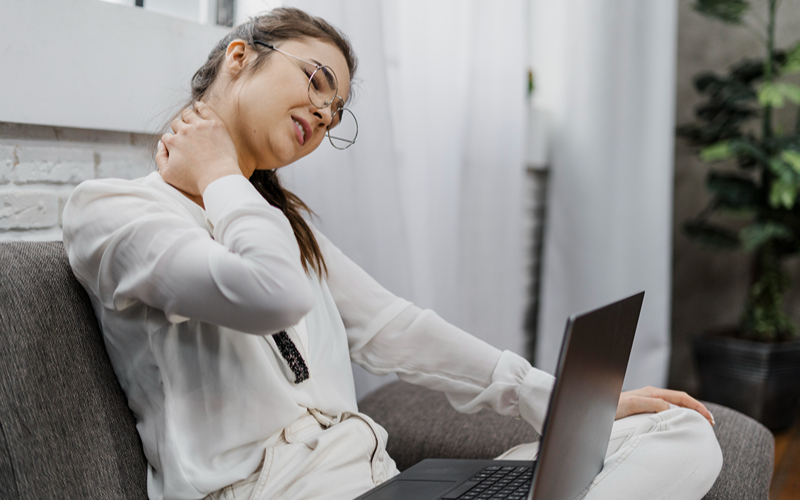 Woman experiencing neck pain while using a laptop on the sofa.